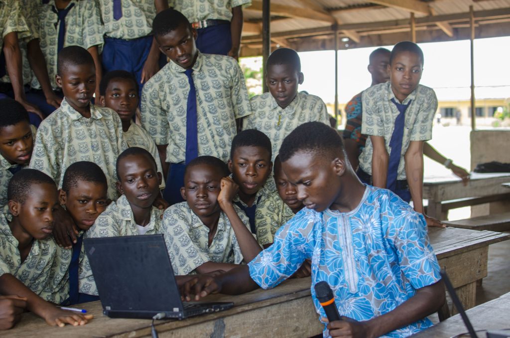 A Tech Now Global facilitator with a microphone helping students at a laptop, TWE School Tour 4.0, October 2021